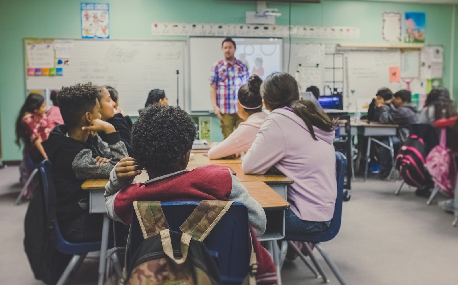 A classroom seen from the back: middle-school students at desks face a teacher in a plaid shirt standing at a whiteboard. The student closest to the camera wears a camouflage backpack.